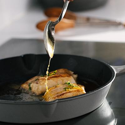 Pan fried sea bass in a cast iron with a spoon dripping butter on top.
